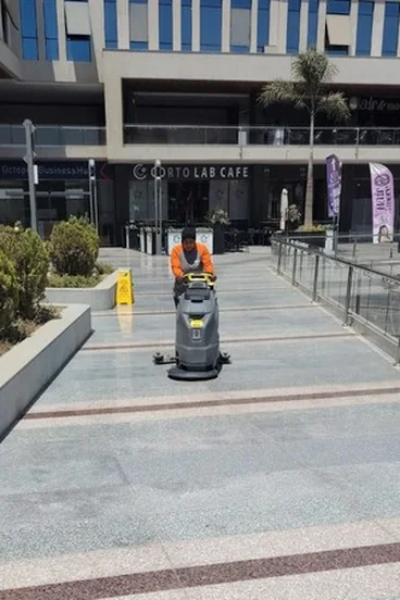 SIAC operator in an orange top driving a floor scrubber across a paved plaza